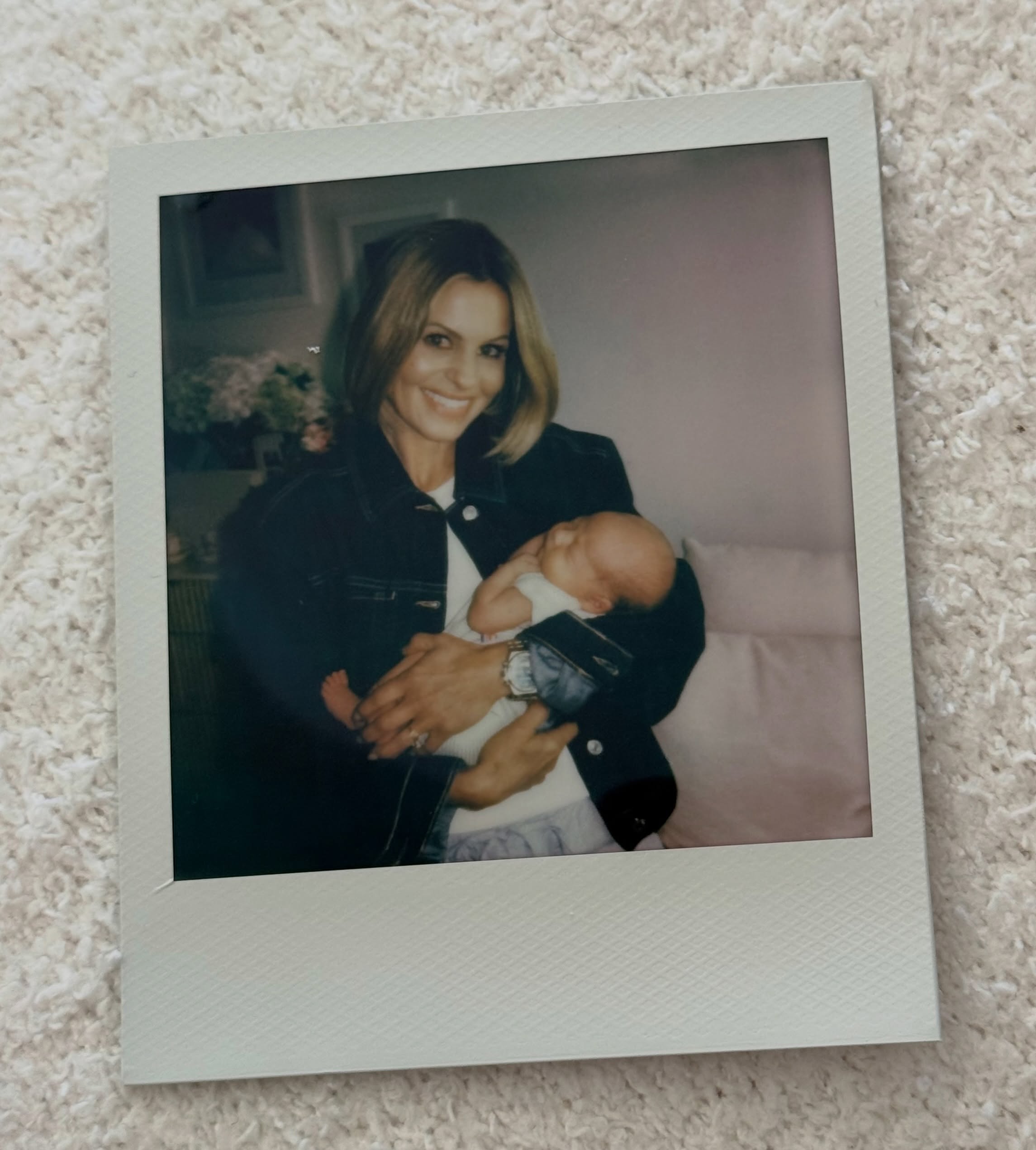 A Polaroid photograph lying on a white textured carpet, showing Candace in a denim jacket smiling while cradling newborn baby Winnie. | Source: Instagram/candacecbure