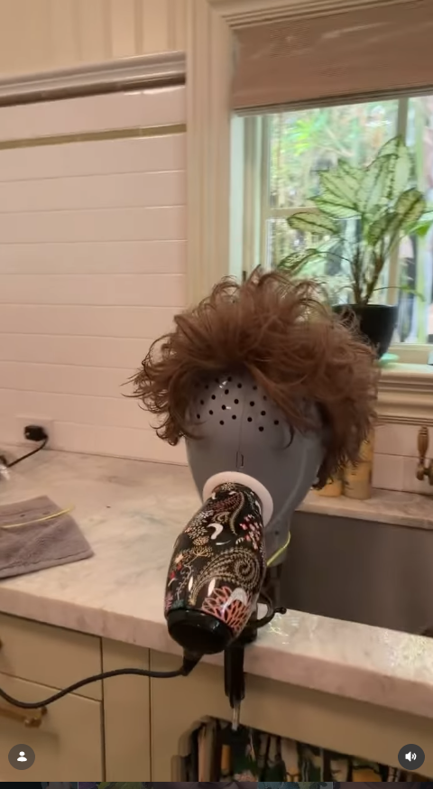 One of McCarthy’s wigs sits on a stand as she uses a hair dryer to dry the freshly washed piece. | Source: Instagram/melissamccarthy