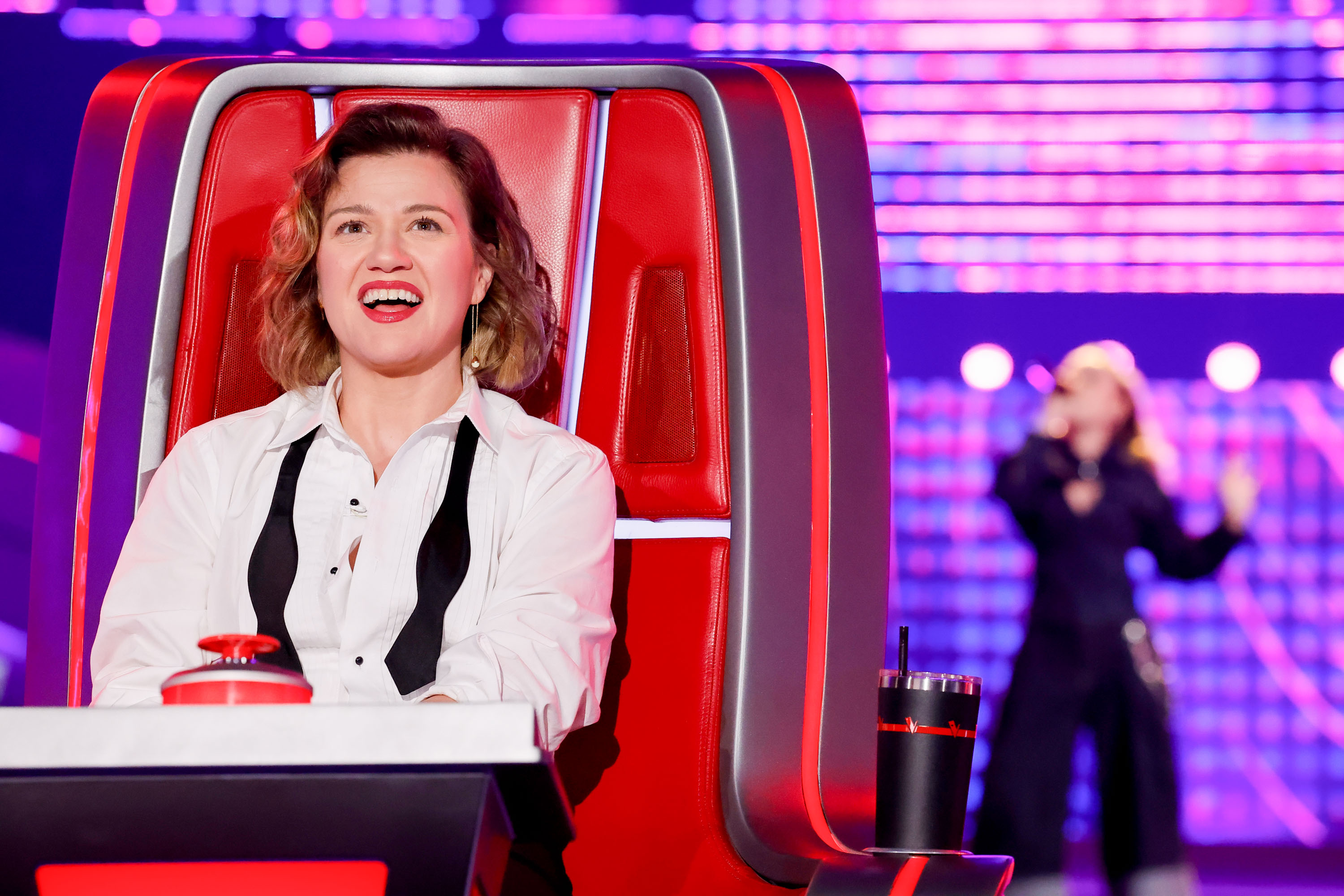 Kelly Clarkson smiled widely from her red coach's chair during "The Blind Auditions Part 3" of "The Voice" Season 29, sporting styled, shoulder-length wavy curls paired with a crisp white button-down shirt and an untied black bow tie drape. | Source: Getty Images
