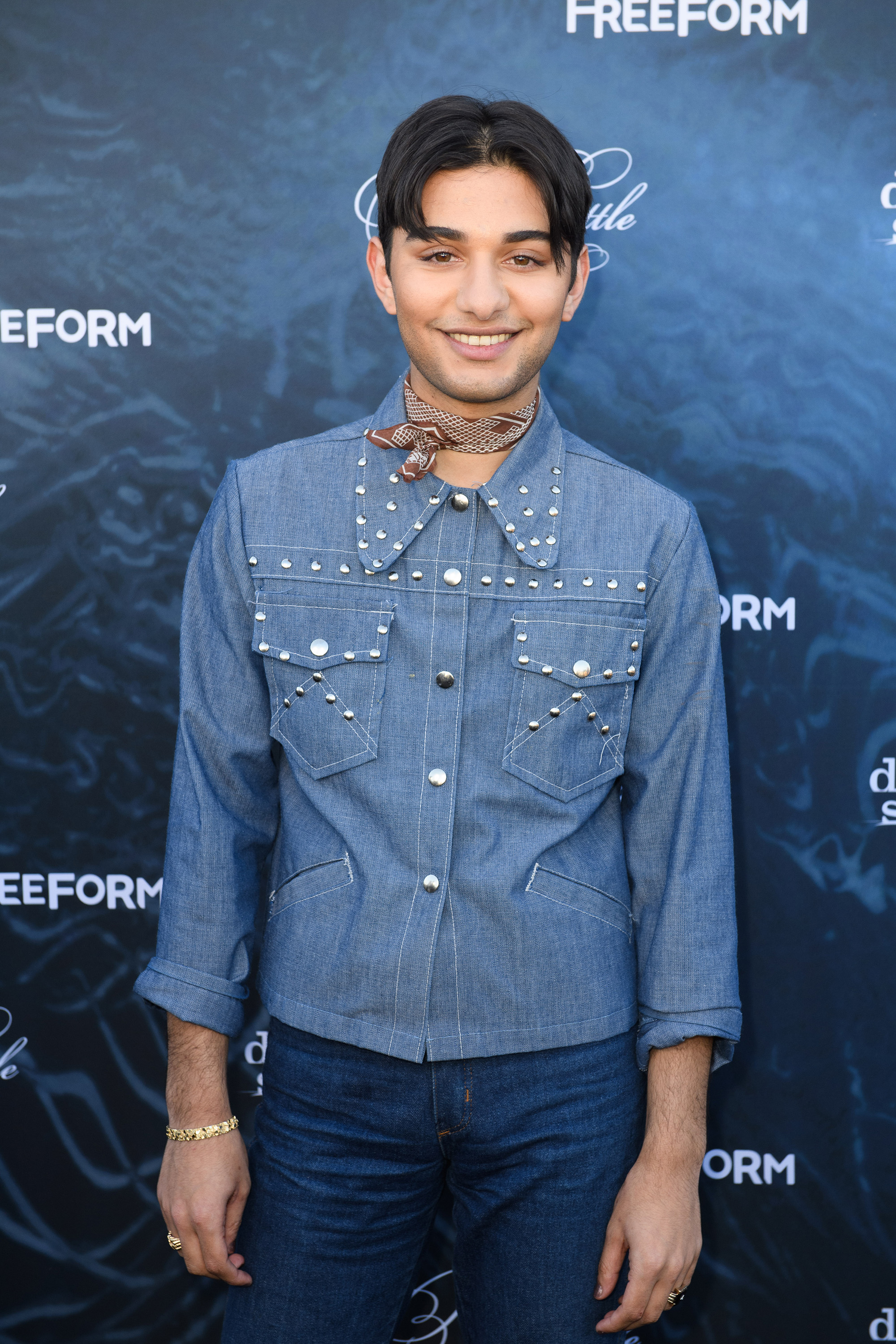 Mark Indelicato attends the premiere event for Freeform's "Dead of Summer" and "Pretty Little Liars" at the Hollywood Forever Cemetery on June 15, 2016, in Los Angeles, California. Channeling a western-inspired aesthetic against the blue Freeform backdrop, he wears a studded light-wash denim shirt with cuffed sleeves and an exaggerated collar, tied with a printed brown neck bandana and paired with dark jeans.