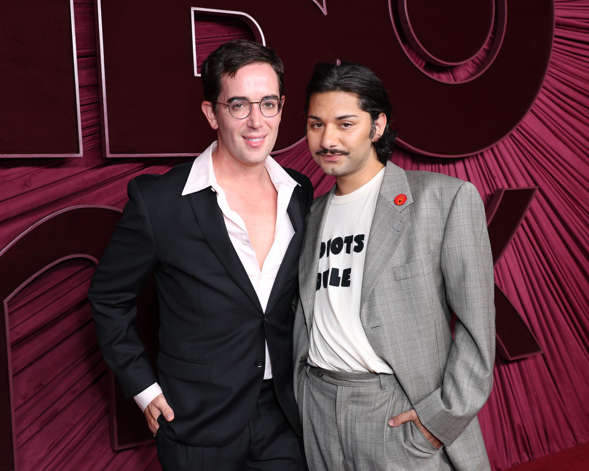 David Watson and Mark Indelicato pose for a full-length red carpet portrait at the HBO Max Primetime Emmy Awards afterparty held at San Vicente Bungalows on September 14, 2025, in Los Angeles, California. Showcasing their evening footwear against the pleated crimson venue backdrop, Watson stands in sleek black dress shoes beside Indelicato, who pairs black-and-white two-tone penny loafers with his relaxed-fit plaid suit and graphic tee.