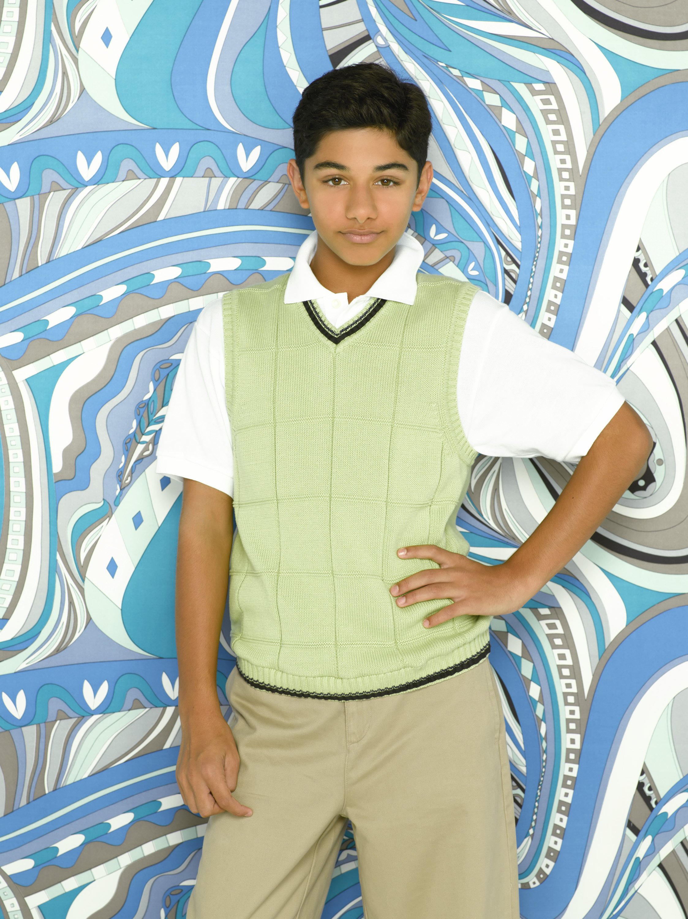 Mark Indelicato stars as Justin Suarez in a promotional portrait for the television series "Ugly Betty," photographed on August 13, 2007. Posing with one hand on his hip against a swirling blue-and-grey geometric patterned background, he wears a light-green knit sweater vest trimmed in black over a crisp white short-sleeved polo shirt, paired with beige trousers.