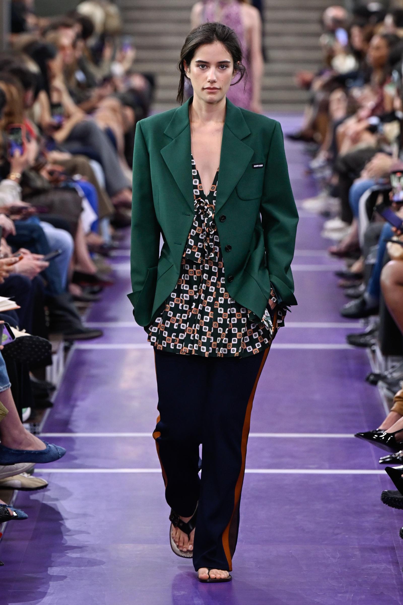Ella Bleu Travolta makes her entrance on the Miu Miu runway in a bold green blazer layered over a patterned top, paired with relaxed navy track pants and black flat sandals. | Source: Getty Images