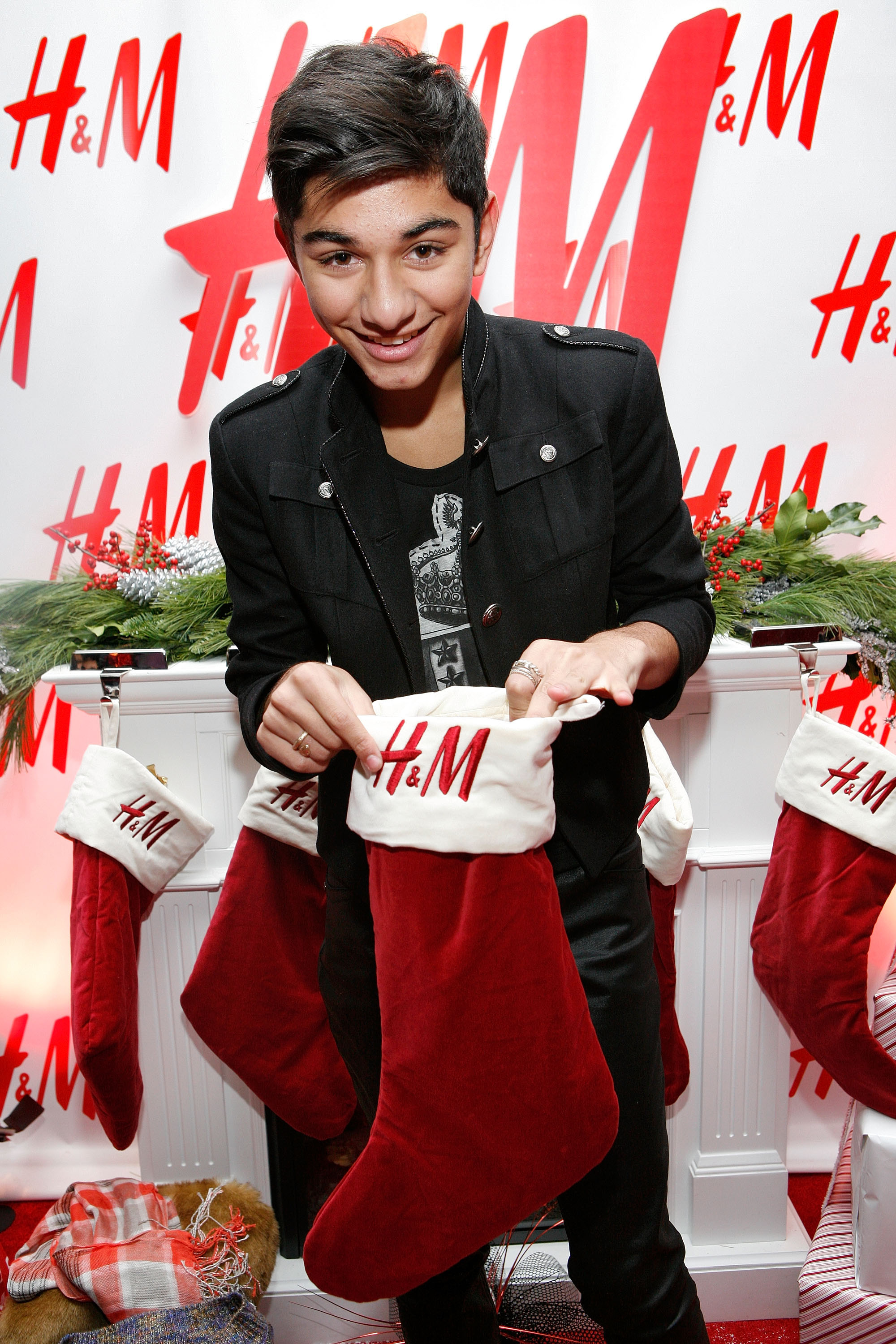 Mark Indelicato smiles playfully as he checks inside a red-and-white embroidered H&M holiday stocking at the H&M Artist Gift Lounge during Z100's Jingle Ball at Madison Square Garden on December 11, 2009, in New York City. He sports a side-swept hairstyle and wears a black military-inspired jacket with silver buttons and flap pockets over a graphic T-shirt, leaning over a decorated faux fireplace mantle lined with holiday stockings and festive greenery.