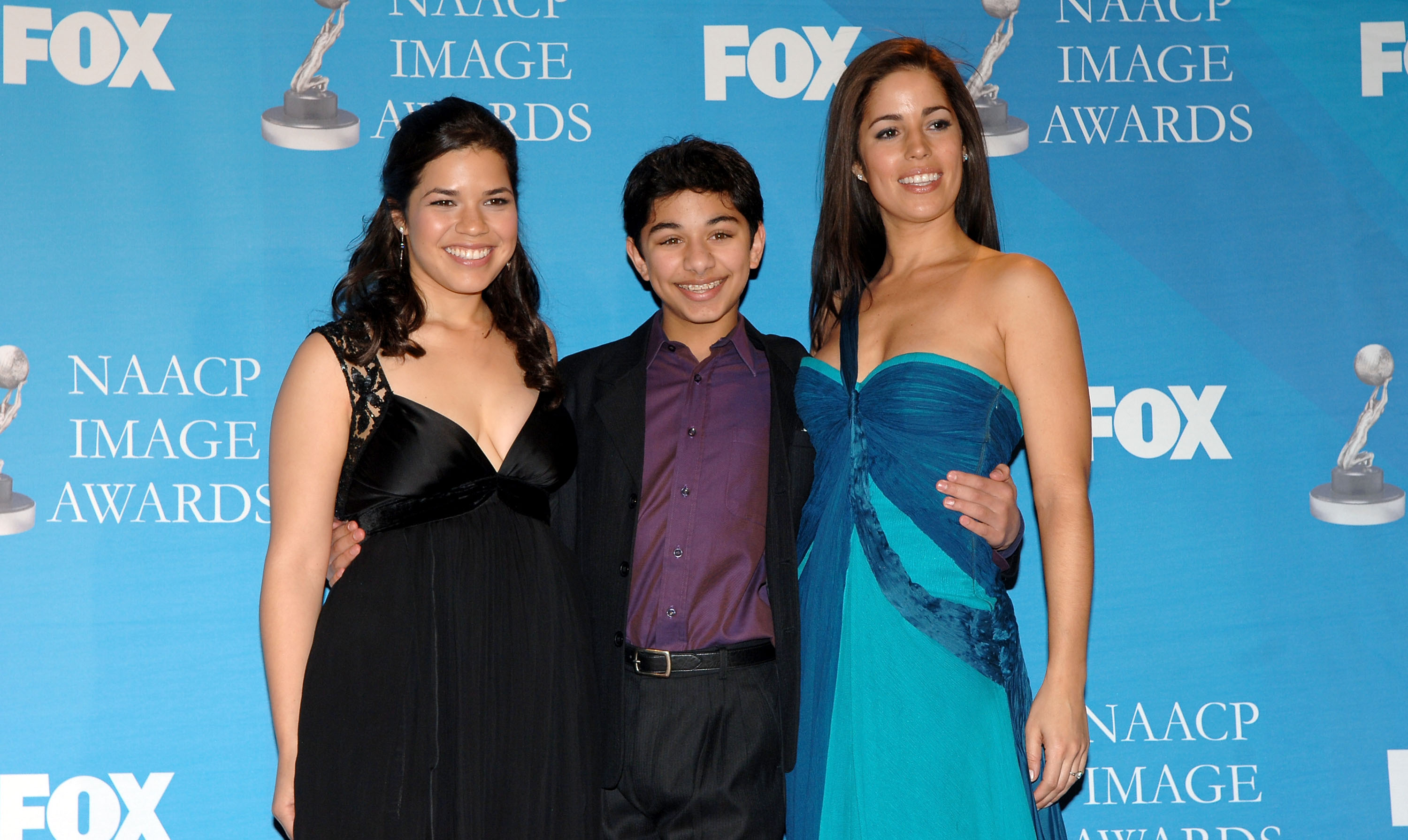 America Ferrera, Mark Indelicato, and Ana Ortiz pose together in the press room celebrating the Outstanding Comedy Series honor for "Ugly Betty" during the 38th Annual NAACP Image Awards at the Shrine Auditorium on March 2, 2007, in Los Angeles, California. Indelicato stands centered in a black suit with a rich purple button-down shirt and belt, flanked by Ferrera in a black empire-waist gown with lace shoulder detailing and Ortiz in a vibrant draped two-tone teal dress against the blue media backdrop.