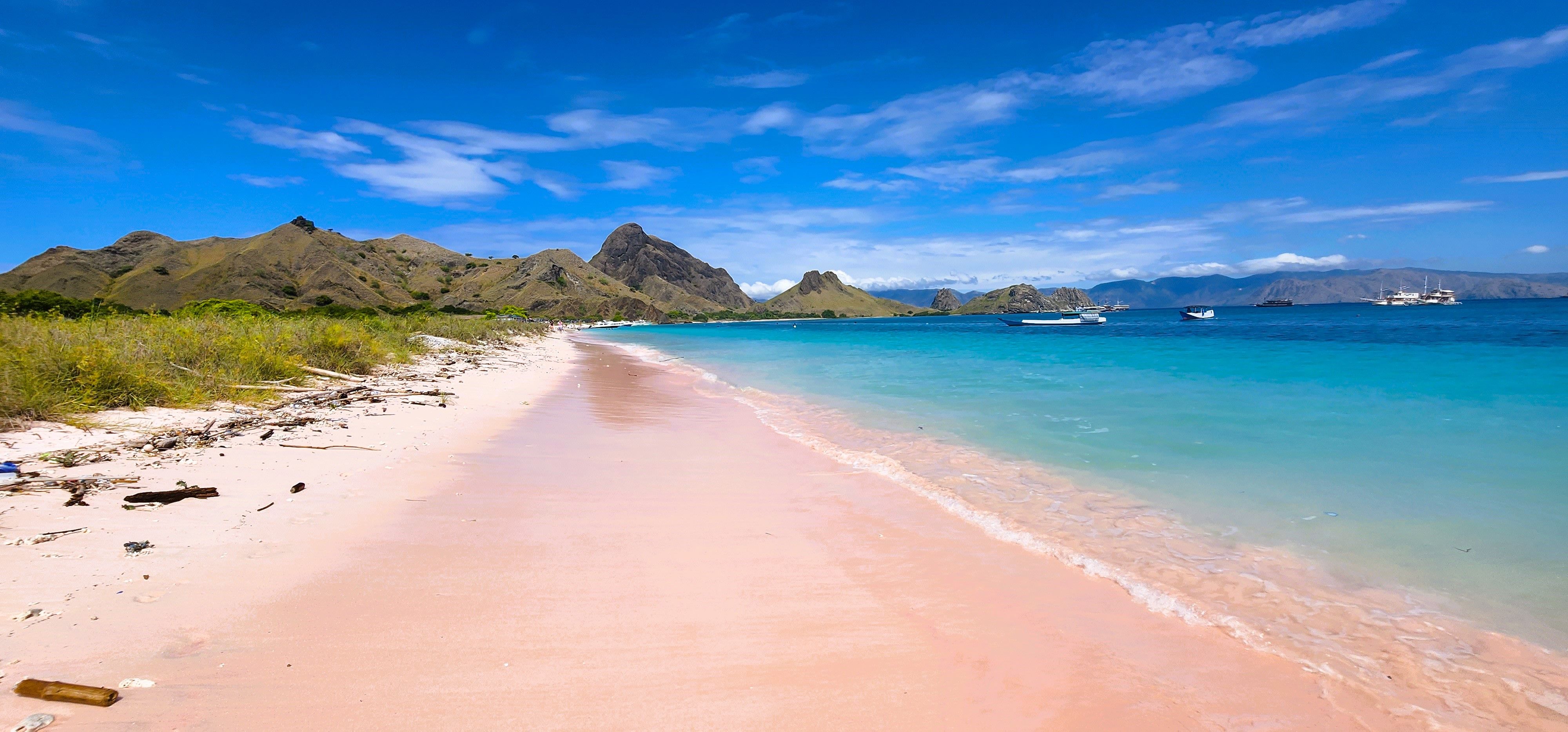 A pink sand beach curves along turquoise water, with hills and boats in the distance. | Source: Pexels