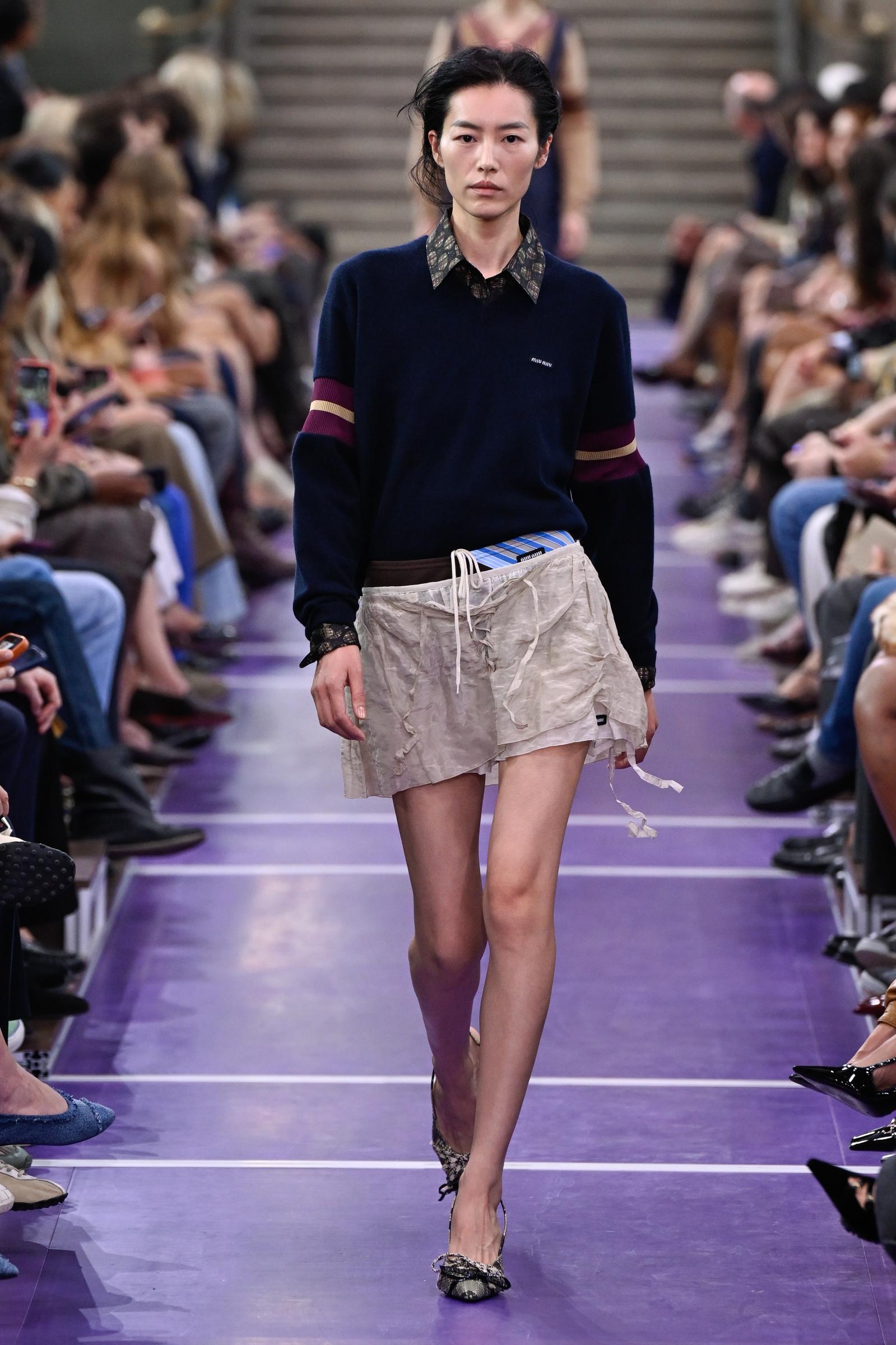 Liu Wen walks the Miu Miu runway in a navy sweater with burgundy and cream sleeve stripes, layered over a patterned shirt and loose cream shorts, with pointed flats completing the look. | Source: Getty Images