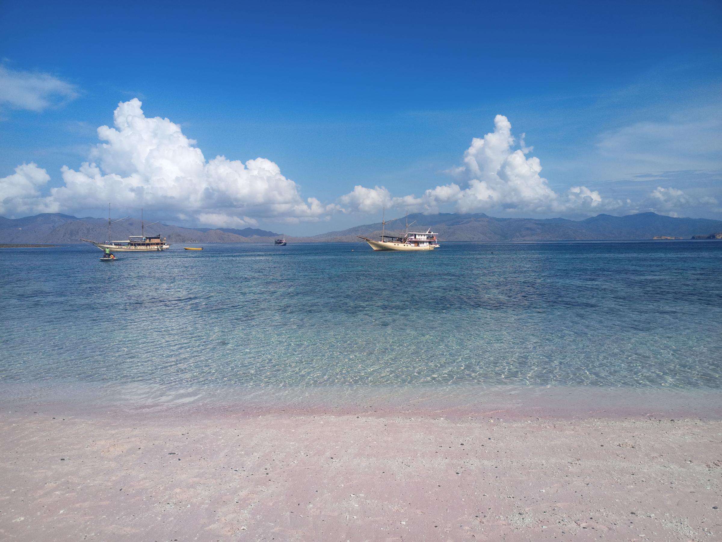 Two boats float in shallow, clear water off a pink sand shore, with mountains in the distance. | Source: Pexels