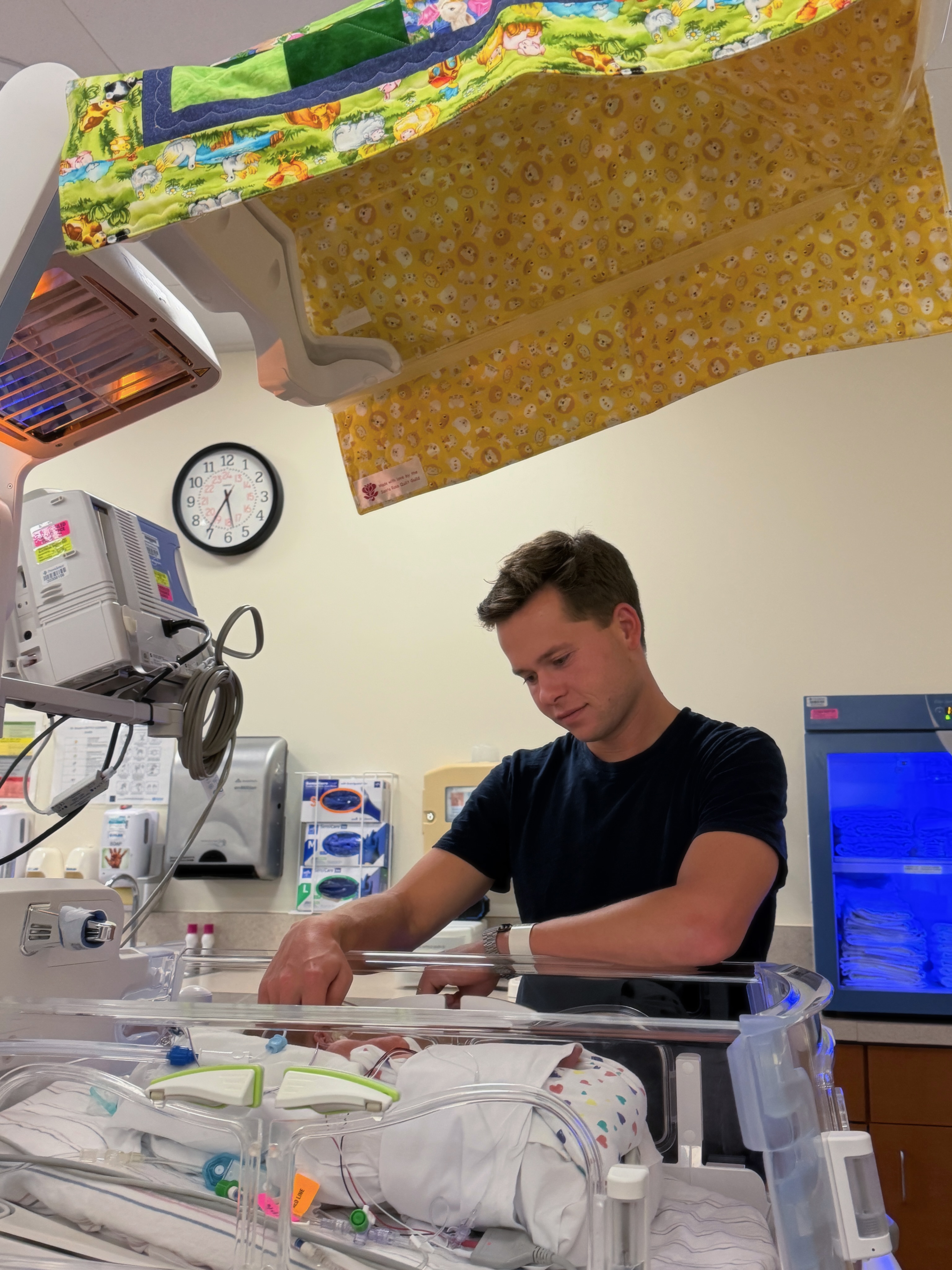 Lev Bure stands beside his newborn son's hospital bassinet, reaching toward Bond as the premature baby receives care surrounded by NICU equipment. | Source: Instagram/elliottpbure
