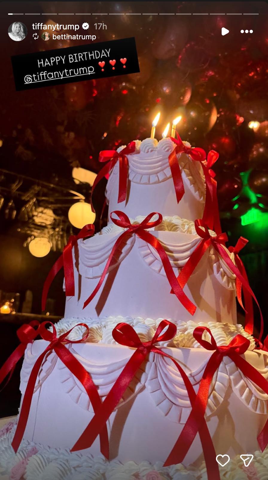 A close-up of Tiffany Trump’s birthday cake shows its elaborate white icing, cascading red satin bows, and lit candles, with festive balloons glowing in the background. | Source: Instagram/tiffanytrump