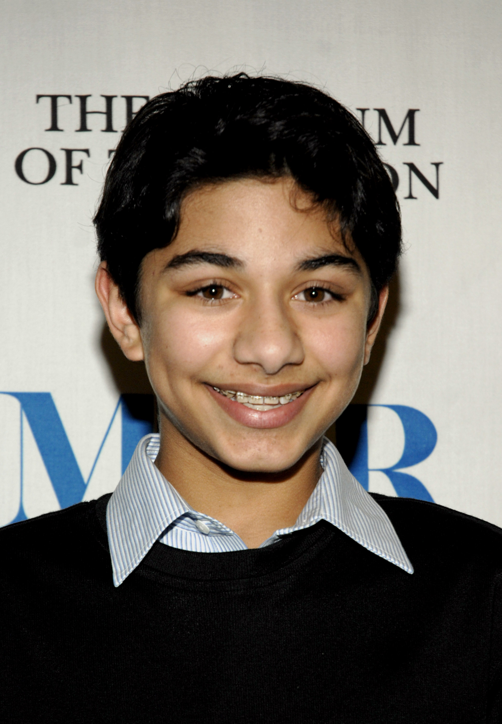 Mark Indelicato smiles in a headshot portrait at the 24th Annual William S. Paley Television Festival presentation of "Ugly Betty" at the Directors Guild of America on March 12, 2007, in Los Angeles, California. He styles a black crewneck sweater over a pale blue-and-white pinstriped collared shirt, showing off his braces and short swept dark hair against the event backdrop.