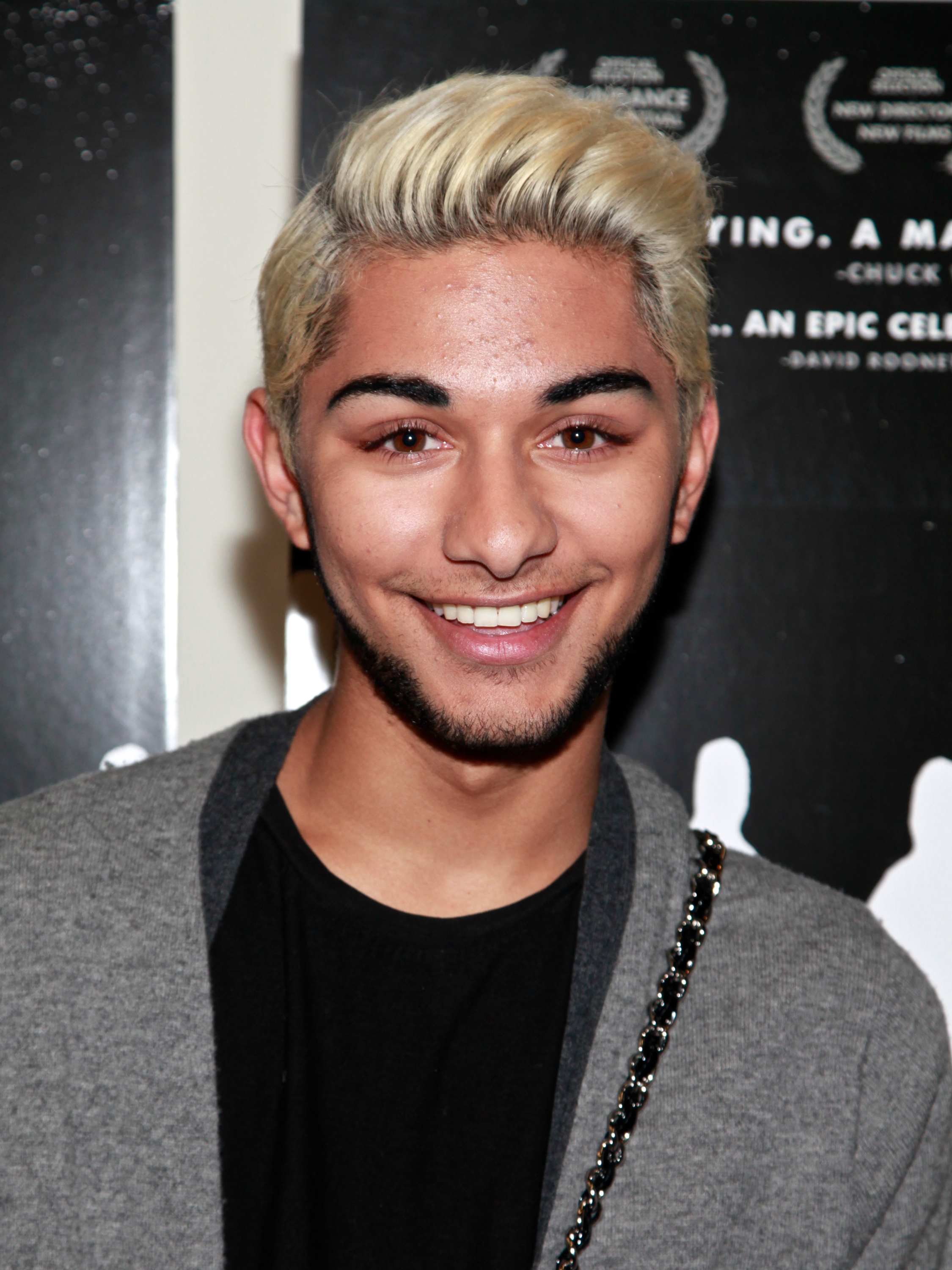 Mark Indelicato attends the New York premiere of "How to Survive a Plague" at the Angelika Film Center on September 19, 2012, in New York City. Showing off platinum-blonde hair contrasted with dark groomed eyebrows and subtle facial hair, he smiles in a close-up portrait wearing a heather-grey knit cardigan over a black crewneck top, accented by a chain-link strap over his shoulder.