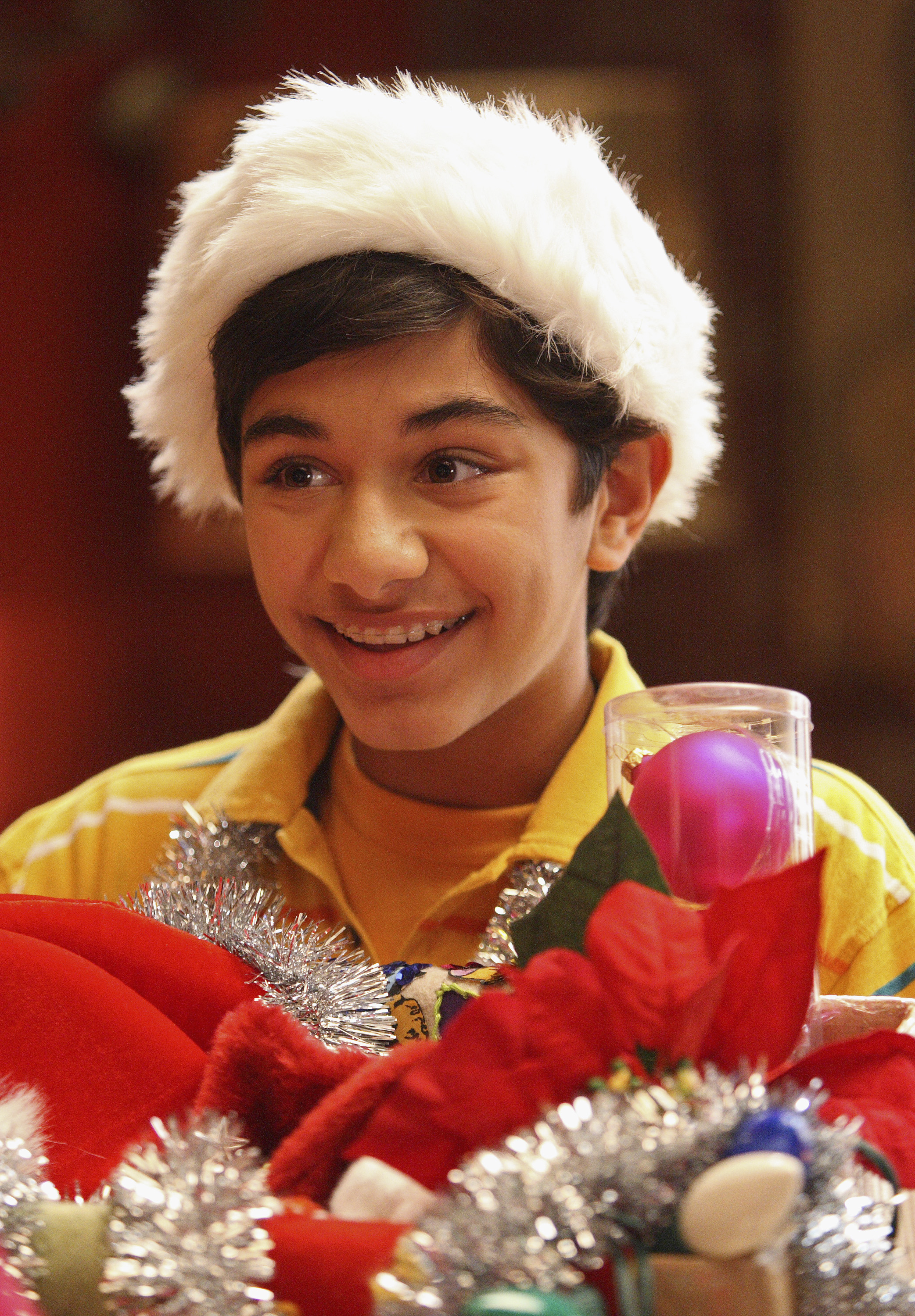 Mark Indelicato portrays Justin Suarez in a scene from the "Giving Up the Ghost" episode of the television series "Ugly Betty," photographed on October 11, 2007. Indelicato smiles in character while wearing a plush Santa Claus hat, a bright yellow collared polo shirt, and silver tinsel draped over his shoulders, surrounded by vibrant holiday decorations, poinsettias, and ornaments.