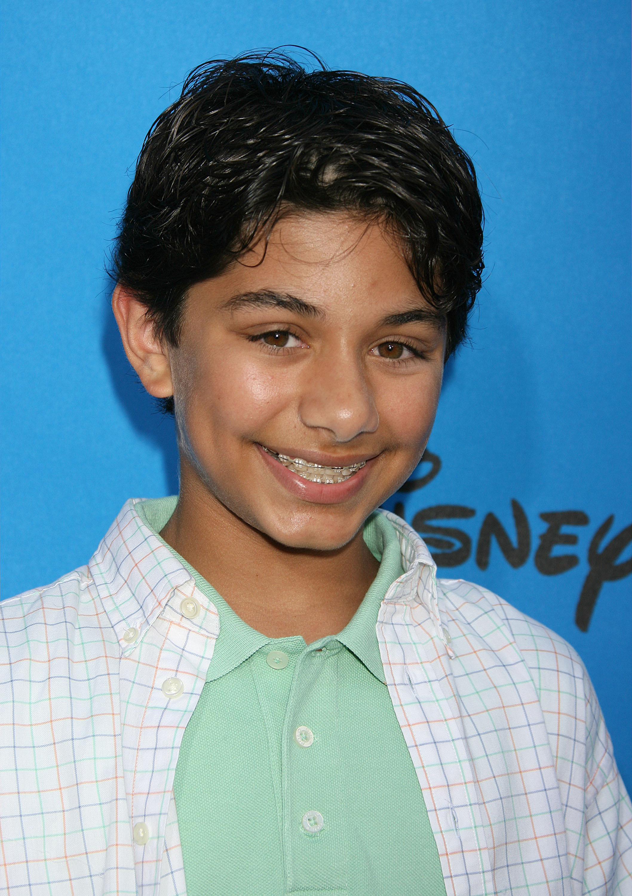 Mark Indelicato smiles in a close-up portrait at the ABC All Star Party arrivals held at the Rose Bowl on July 19, 2006, in Pasadena, California. Sporting short wavy dark hair and braces, he wears a light mint-green polo shirt beneath an open white short-sleeved button-down featuring a pastel windowpane check pattern, set against a bright blue Disney step-and-repeat backdrop.