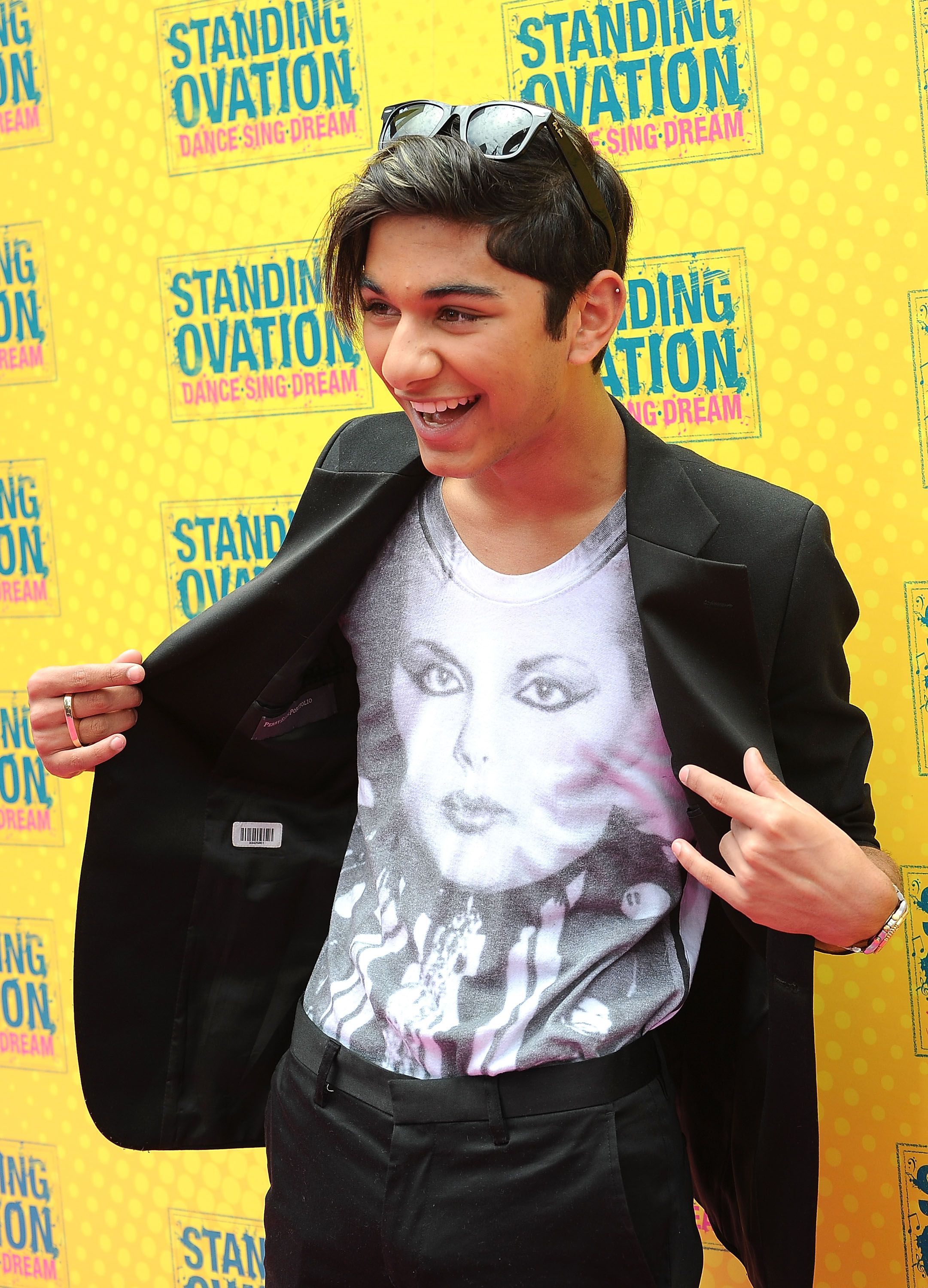Mark Indelicato arrives at the Los Angeles premiere of "Standing Ovation" at Universal CityWalk on July 10, 2010, in Universal City, California. Flaunting a bold, expressive style, he holds open his tailored black blazer to highlight a white graphic tee featuring a portrait of Liza Minnelli, completing the look with black trousers, sunglasses resting atop his side-swept hair, and a playful hand gesture against the vibrant yellow step-and-repeat backdrop.