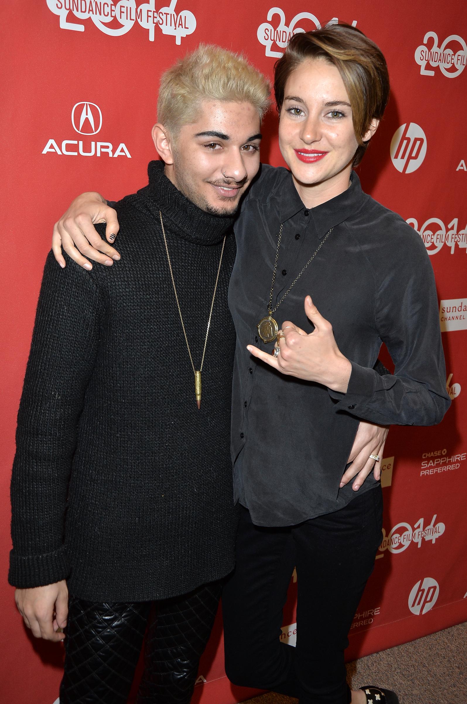 Mark Indelicato and co-star Shailene Woodley pose arm-in-arm at the premiere of "White Bird in a Blizzard" at Eccles Center Theatre during the 2014 Sundance Film Festival on January 20, 2014, in Park City, Utah. Indelicato sports bleached blonde hair, a black knit turtleneck sweater, quilted leather trousers, and a bullet pendant chain, smiling beside Woodley, who rocks a short pixie cut, dark button-down shirt, black pants, and a playful hand gesture against the red festival backdrop.