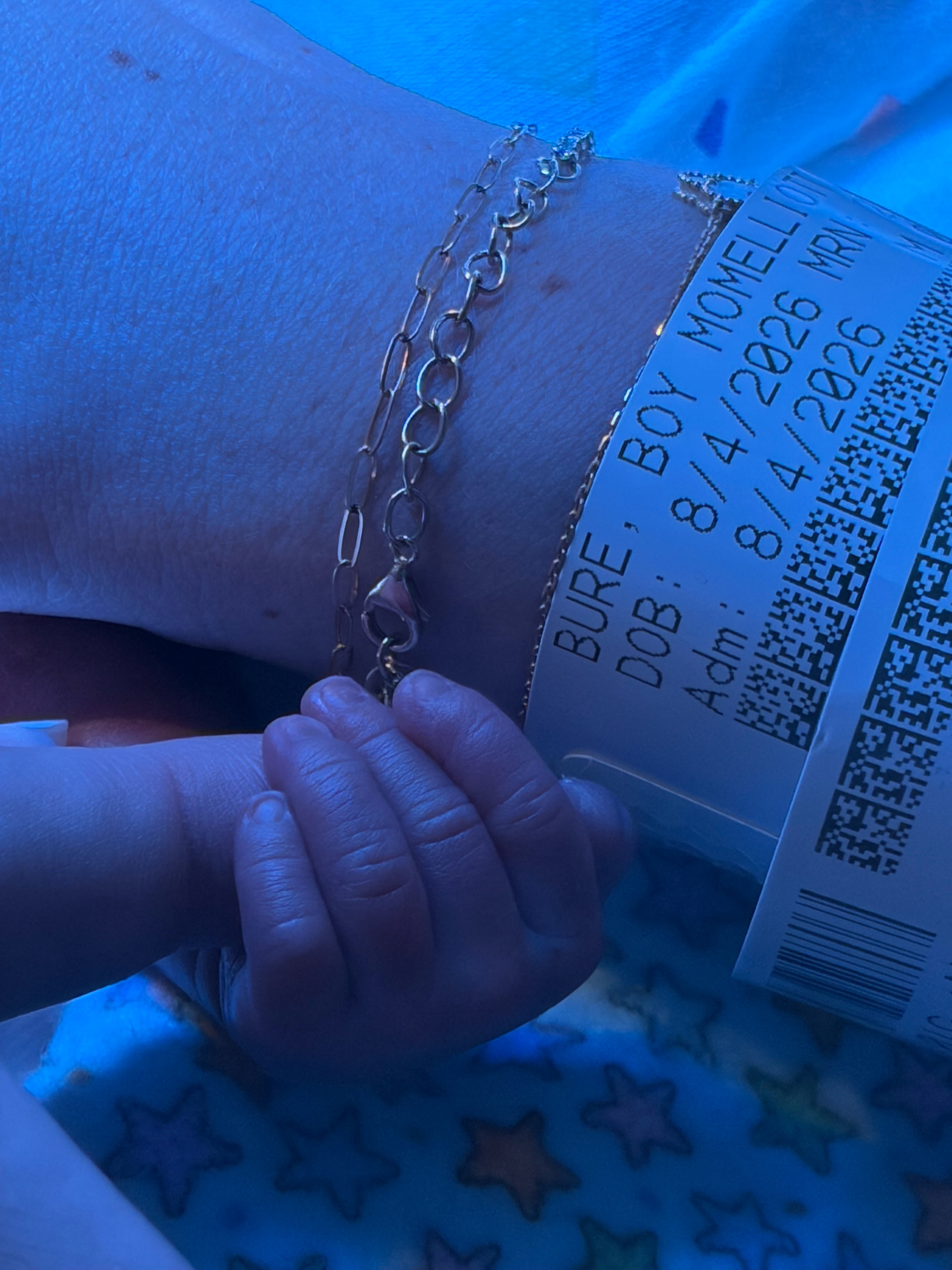 Newborn Bond curls his fingers around his mother's bracelet beside a hospital wristband identifying Elliott Bure and displaying his August 4, 2026, birth date.