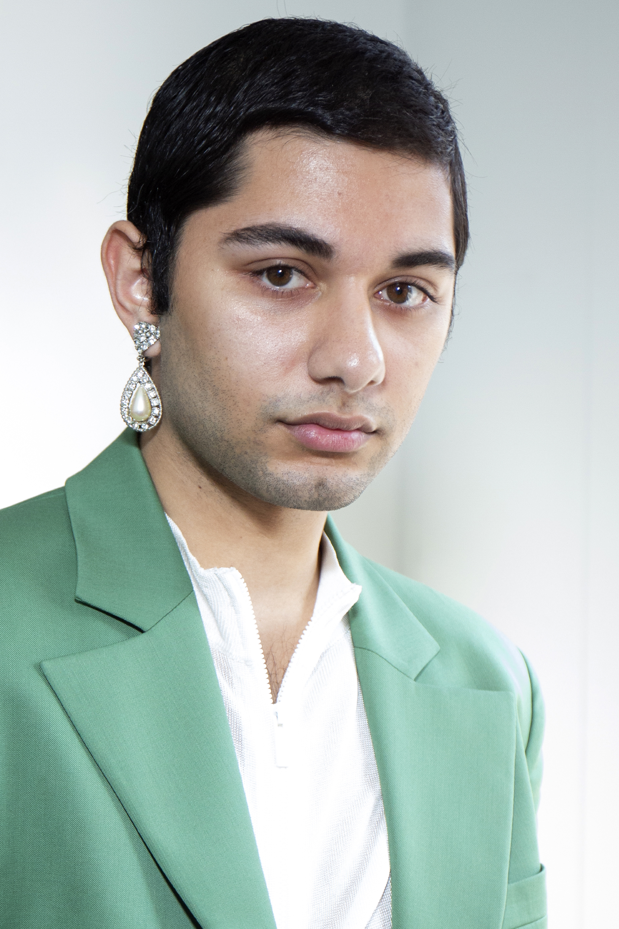 Mark Indelicato delivers a poised, close-up pose attending the "Wan Hung" presentation at Pier 59 Studios on February 5, 2019, in New York City. Making a high-fashion statement, he pairs an emerald green tailored blazer over a white zip-up inner layer, accessorized with a striking crystal-and-pearl teardrop chandelier earring and subtle five o'clock shadow.