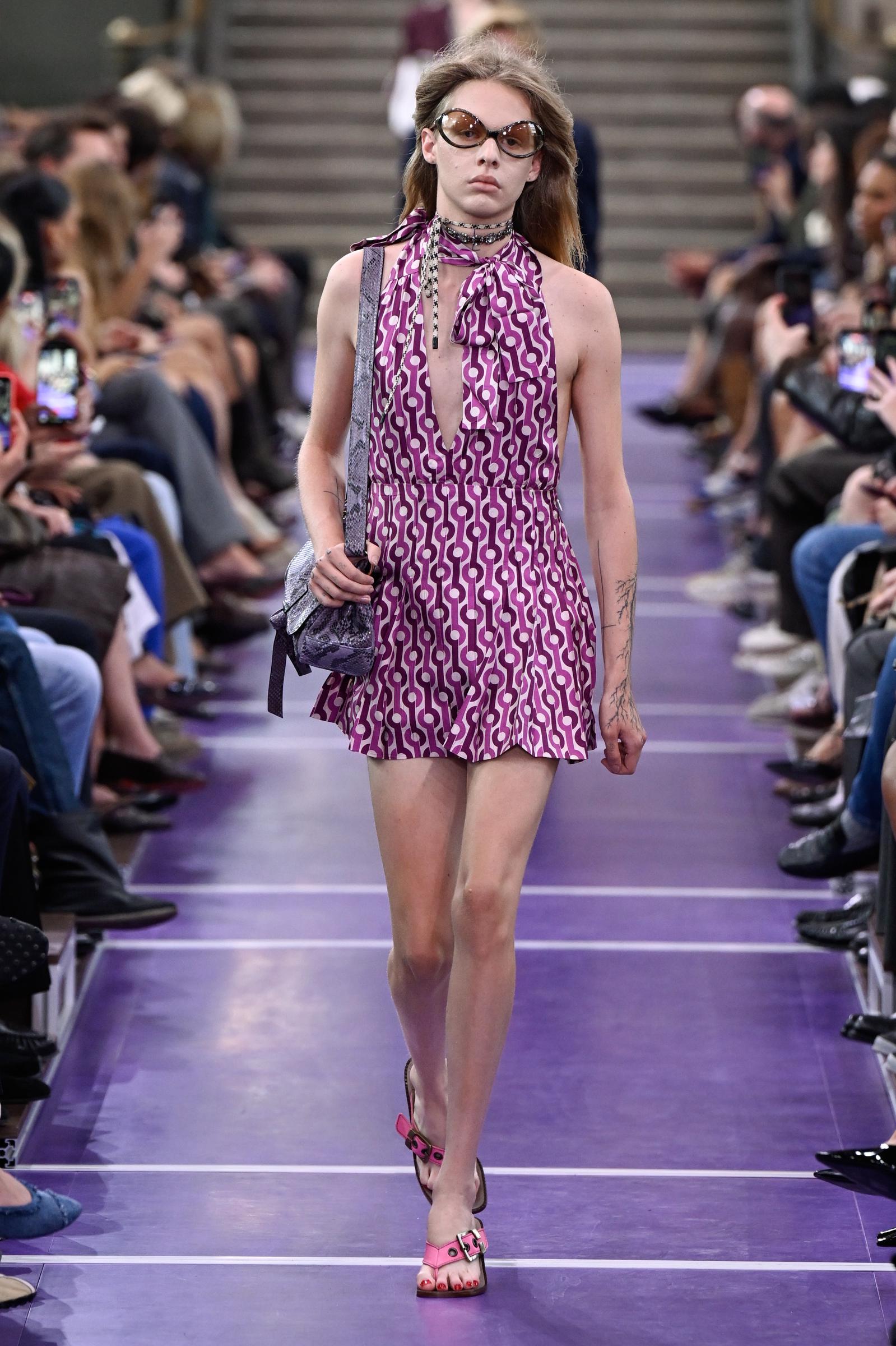A model closes out the runway sequence in a purple-and-white geometric halter mini dress, oversized sunglasses and layered chokers, carrying a metallic shoulder bag and wearing pink strappy heels. | Source: Getty Images