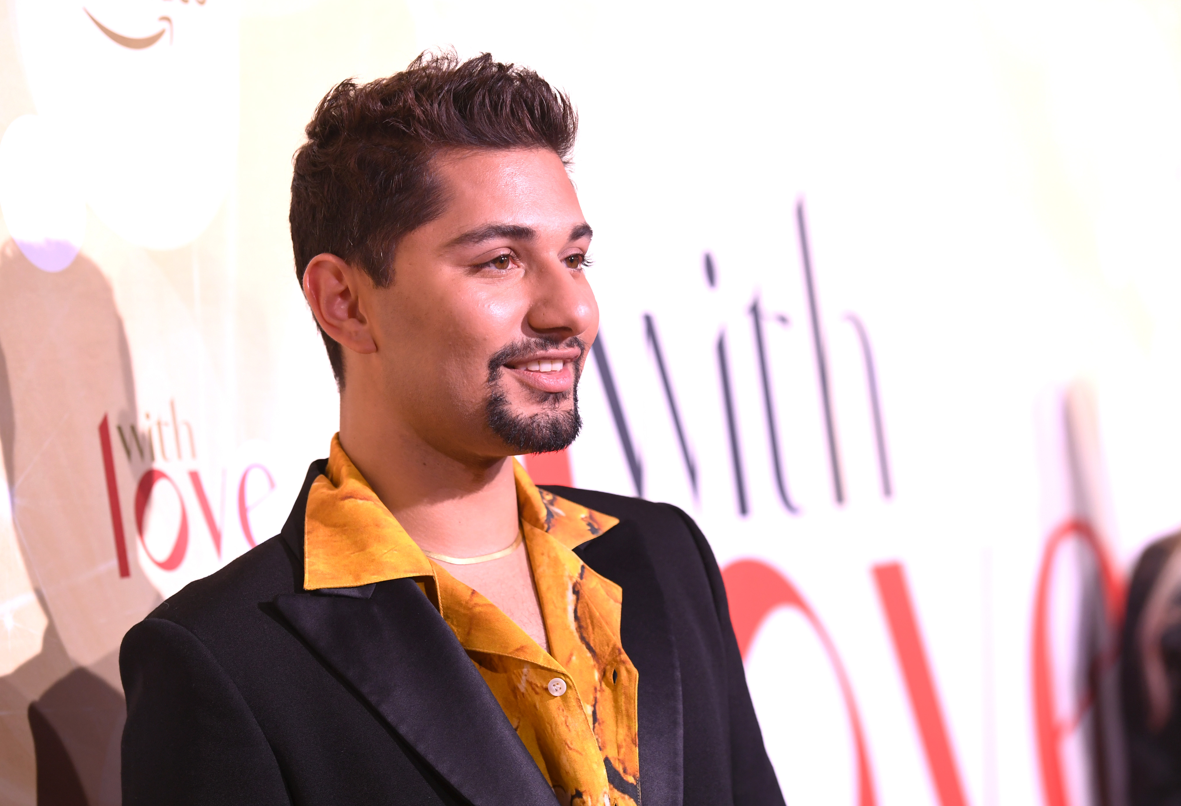 Mark Indelicato smiles in profile at the Prime Video red carpet premiere for the romantic comedy holiday series "With Love" at NeueHouse Los Angeles on December 9, 2021, in Hollywood, California. Sporting a neatly trimmed goatee and styled dark hair, he steps out in a black tuxedo jacket with satin lapels layered over an open-collar golden-orange patterned shirt, accented with a delicate gold chain necklace.
