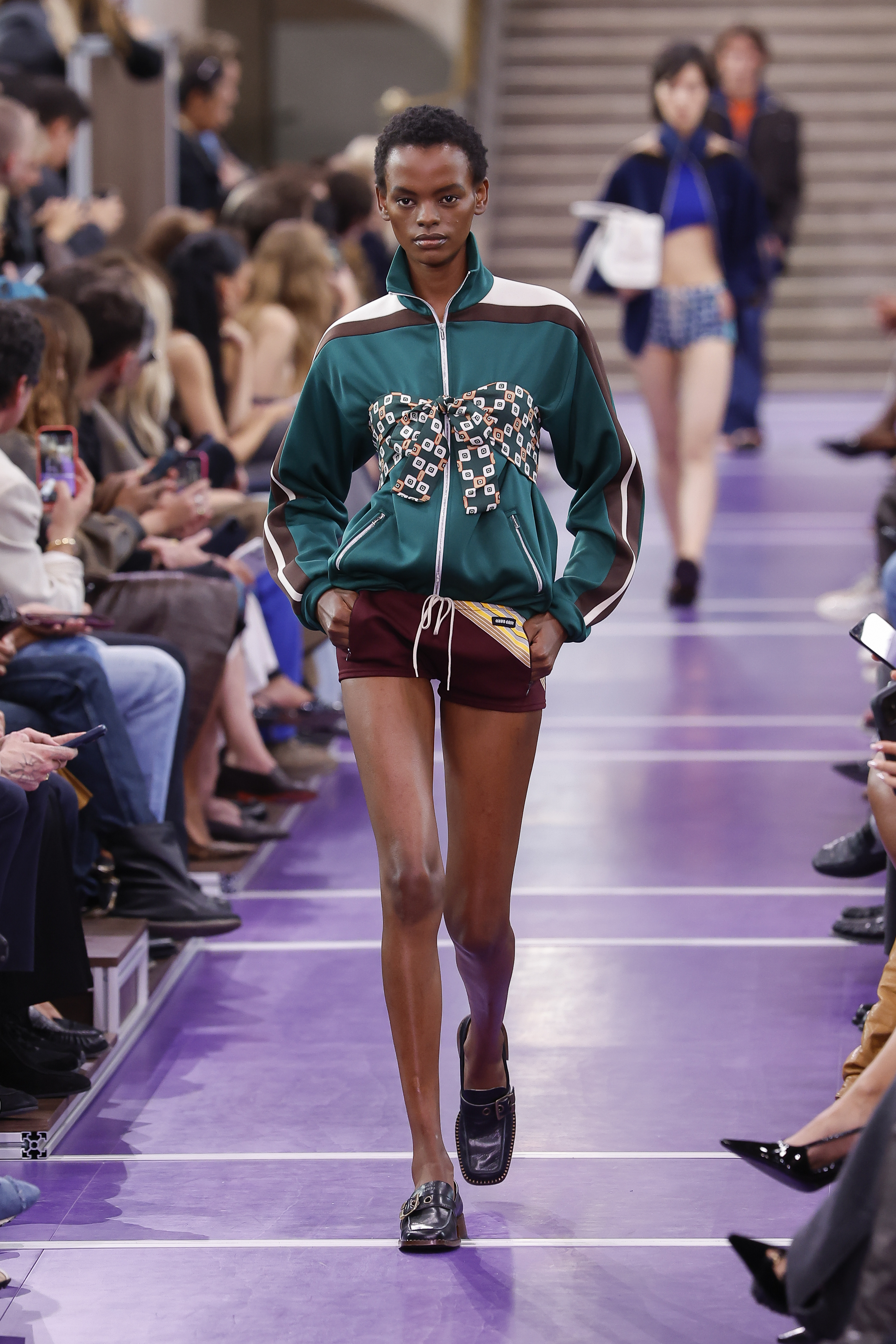 A model walks the Miu Miu runway in a sporty teal track jacket with racing-inspired stripes and a patterned bow detail, paired with burgundy shorts and black loafers. | Source: Getty Images
