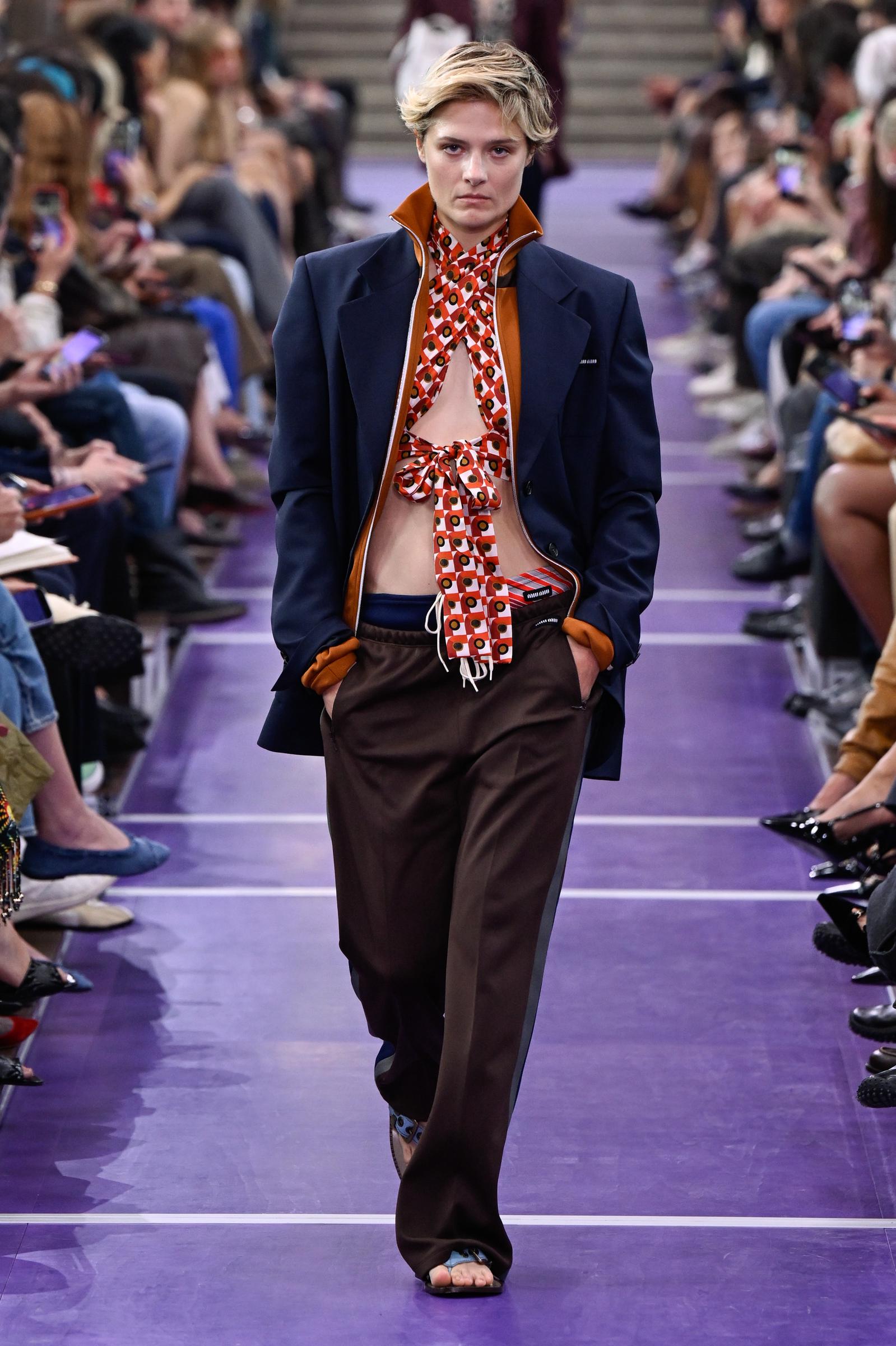 Louisa Jacobson walks the Miu Miu runway in a tailored navy blazer with contrasting orange details over a bold patterned halter top, paired with loose brown trousers and flat open-toe sandals. | Source: Getty Images