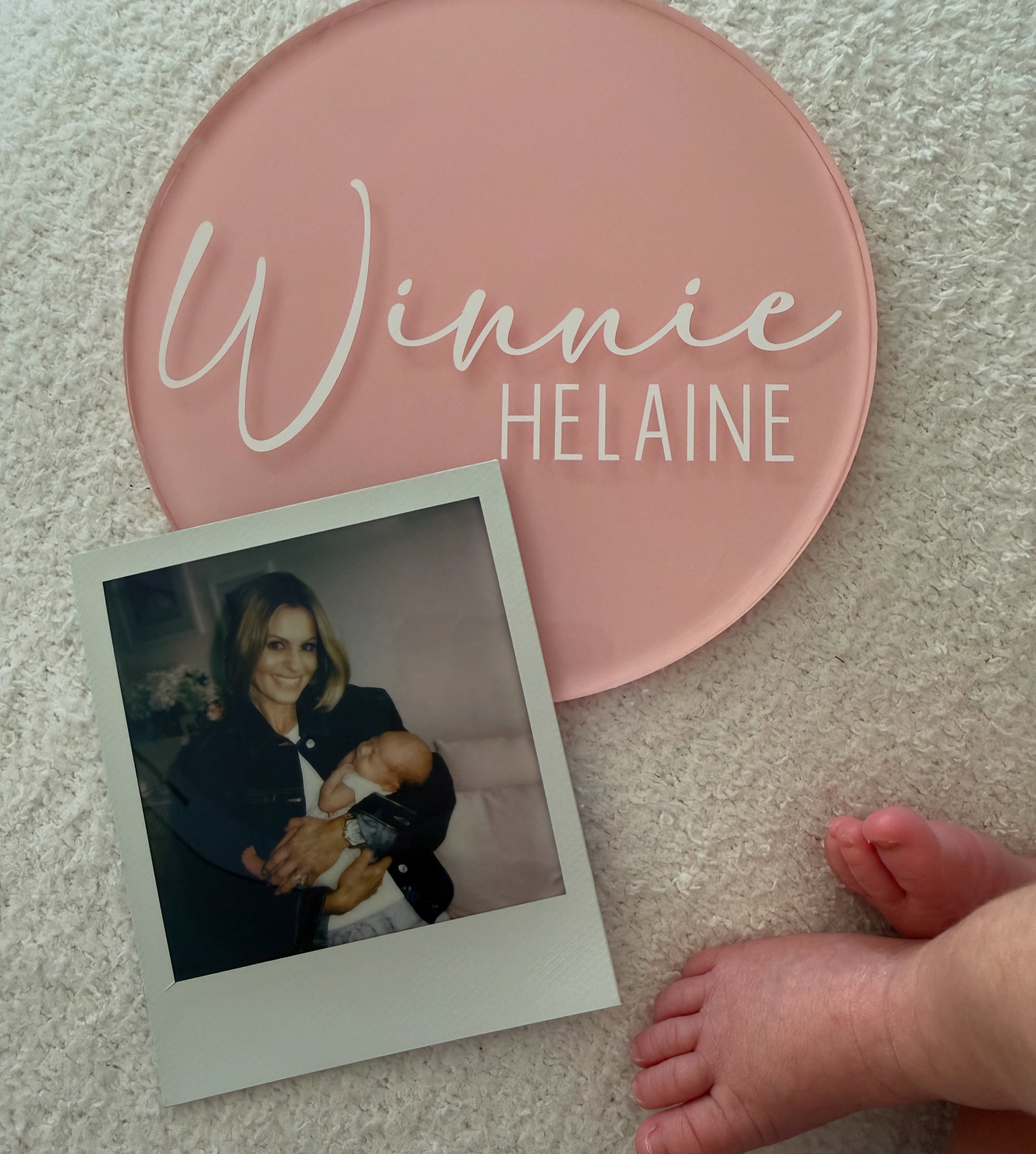 A top-down photo featuring a pink circular acrylic sign bearing the baby's nae, the Polaroid of Candace and Winnie, and baby Winnie's tiny feet in the bottom corner. | Source: Instagram/candacecbure