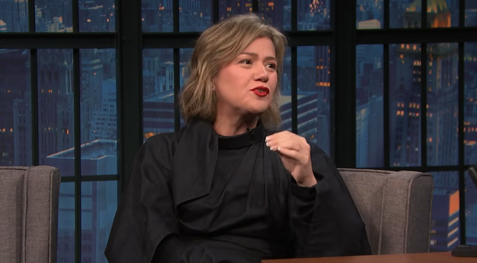 Sitting down on "Late Night with Seth Meyers" on September 21, 2026, Kelly Clarkson gestured with her hand as she hilariously recounted the viral clip from "The Voice" where she offered her own clip-in hair extension to a contestant, debuting her chic new short bob paired with bold red lipstick and a high-neck black dress. | Source: YouTube/Late Night with Seth Meyers