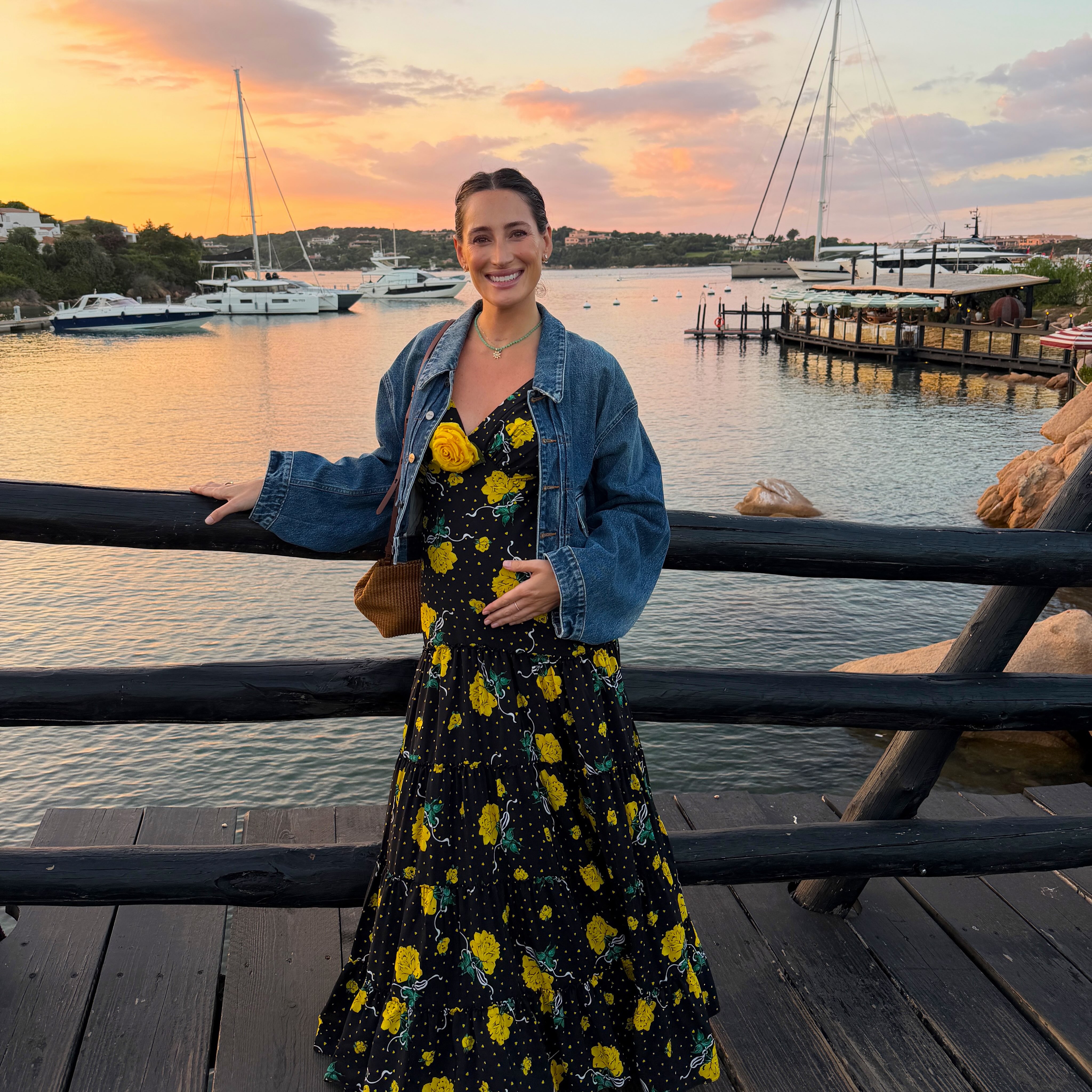 Jessica Springsteen cradles her baby bump in a floral dress and denim jacket while posing beside the water at sunset. | Source: Instagram/jessicaspringsteen